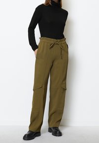 Zelenohnědé cargo kalhoty s elastickým pasem, stahovací šňůrou a bočními kapsami. Spárovány s obepnutým černým rolákem a černými botami.