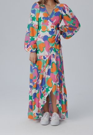 Femme portant une longue robe portefeuille colorée avec des motifs de cœurs et de formes abstraites, associée à des baskets blanches, debout devant un fond uni.