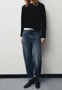 Pull noir en tissu duveteux associé à un jean en denim à lavage clair et coupe ample, agrémenté d'une ceinture noire. Porté avec des bottines noires.