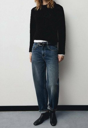 Pull noir en tissu duveteux associé à un jean en denim à lavage clair et coupe ample, agrémenté d'une ceinture noire. Porté avec des bottines noires.