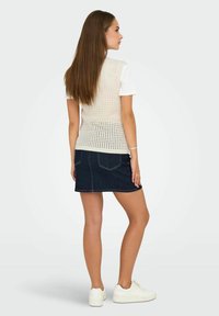 Top senza maniche a maglia color crema con motivo aperto abbinato a una gonna in denim scuro. La modella indossa sneakers bianche, mostrando un outfit casual da dietro.