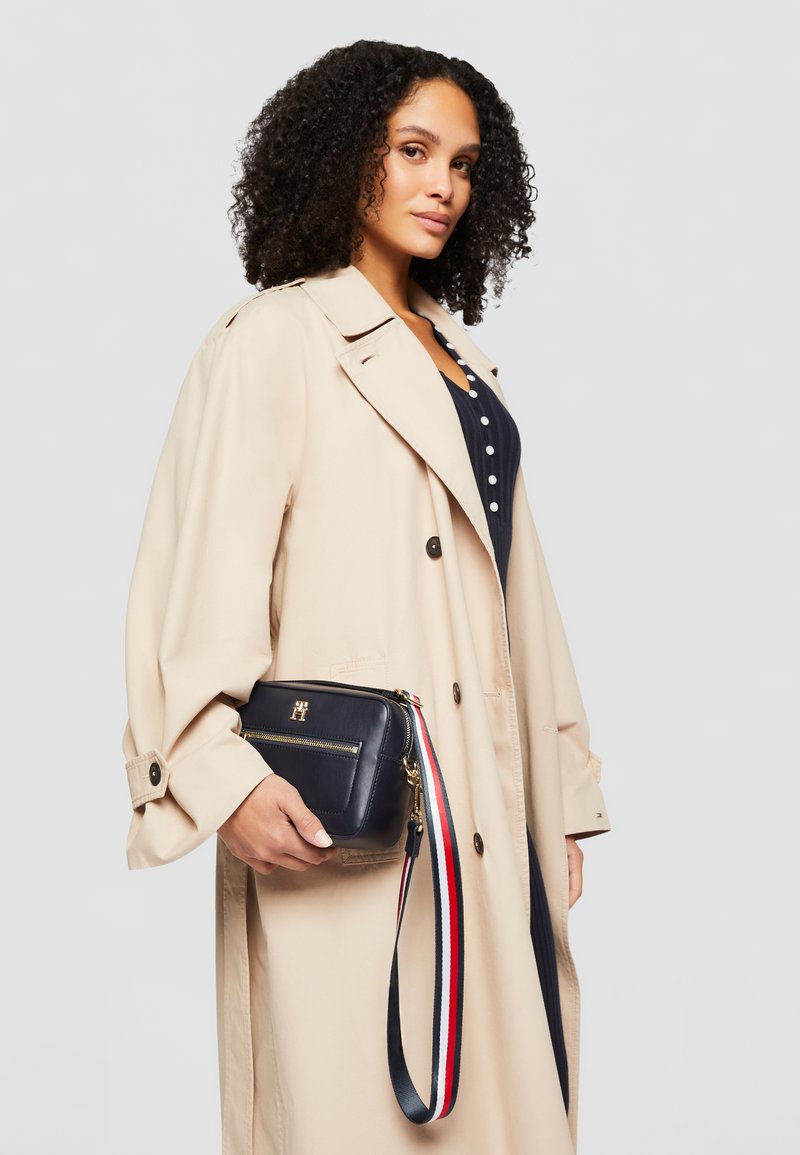 Borsa a tracolla in pelle blu navy con zip dorata, tracolla a righe rosse e bianche e un accento con logo. Il modello indossa un trench coat beige.
