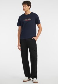 Tamsiai mėlynas marškinėlis su baltu ir raudonu "GUESS JEANS" logotipu, derinamas su juodomis krovinių kelnėmis, turinčiomis daugybę kišenių, ir smėlio spalvos sportbačiais.
