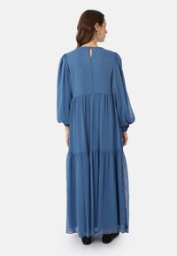 Long blue dress made of flowy fabric, featuring puffed sleeves, a tiered skirt, and a back keyhole detail. Black shoes visible.