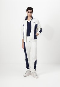 Veste zippée blanche avec des accents bleu marine, pantalon ajusté assorti et t-shirt bleu marine à col en V. Les détails incluent un logo et un design sportif.