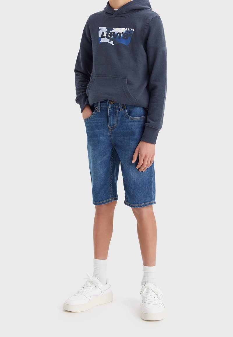 Mörkblå hoodie med en vit och blå Levi's logotyp, i kombination med ljusblå denimshorts och vita sneakers, med en slät textur.