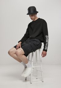 Tânăr purtând pălărie bucket neagră, cămașă neagră cu mâneci lungi și text alb pe mânecă, pantaloni scurți cargo negri, șosete albe și adidași albi, stând pe un scaun alb.