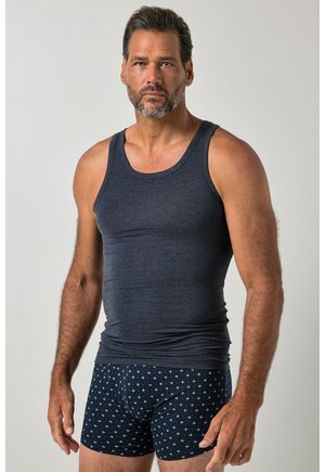 Hombre con cabello corto y oscuro y barba, que lleva una camiseta sin mangas gris oscuro y calzoncillos bóxer azul marino con patrones de cruces blancas.