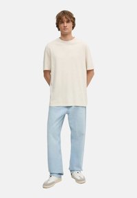 Camiseta de algodón color beige con cuello redondo, combinada con jeans rectos de color azul claro y zapatillas blancas con detalles en gris.