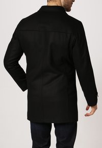 Cappotto di lana nera con taglio dritto, dotato di un colletto rialzato e dettagli di cucitura minimi sul retro. Tessuto morbido e senza chiusure visibili.