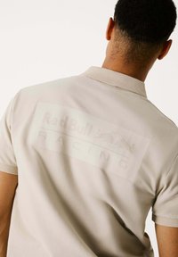 Šviesiai ruda polo marškinėliai su apykakle ir trumpomis rankovėmis, išsiskiriantys dideliu toniniu "Red Bull Racing" logotipu ant nugaros. Lygus paviršius.