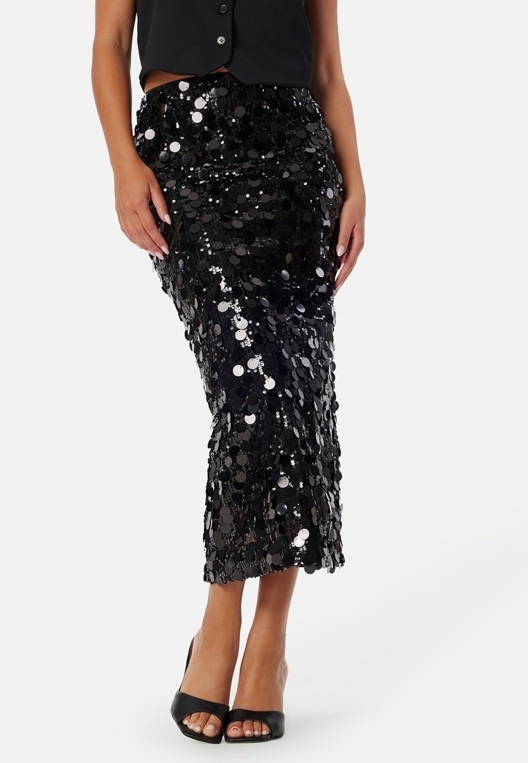 Femme portant une jupe midi noire à sequins associée à des chaussures noires à talons hauts et bouts ouverts, ainsi qu'un gilet noir sans manches.