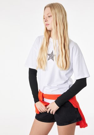 Lyshåret kvinde iført en hvid T-shirt med stjerner printet ovenpå sorte langærmede, sorte shorts og en rød sweater bundet om taljen.