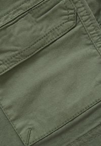 Olijfgroene katoenen stof met een gestructureerd oppervlak, opvallende stiksels en een opgestikte zak. Soepele, duurzame textuur met een matte afwerking.