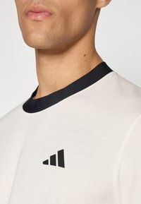 Bela majica s črnim rebrastim ovratnikom in črnim Adidasovim logotipom na levi strani. Tkanina se zdi gladka in lahka.
