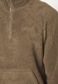 Brauner Fleece-Pullover mit halbhohem Reißverschluss, strukturierter Oberfläche und einem dezenten Logo auf der Vorderseite. Weiches, warmes Material mit langen Ärmeln.