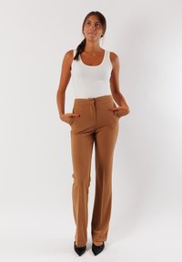 Pantaloni marroni su misura con vita alta, pieghe frontali e due tasche laterali, abbinati a un top bianco aderente e tacchi neri.