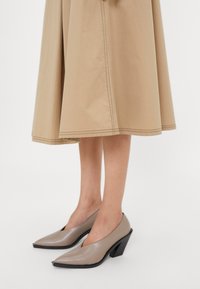 Jambes portant des talons beiges pointus avec des talons blocs inclinés, associés à une jupe midi beige avec des détails de couture visibles.