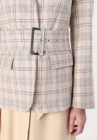 Blazer à carreaux beige avec une ceinture à boucle rectangulaire métallique, accents bleus subtils, tissu texturé et poches latérales. Coupe ajustée.