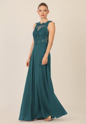 Teal evening gown with intricate beaded bodice and sheer neckline. Flowing fabric drapes elegantly to the floor, complemented by strappy heels.