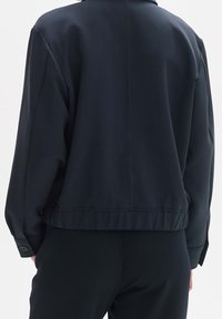 Granatowa kurtka typu bomber o luźnym kroju, z kołnierzem, elastycznym pasem i mankietami na guziki. Gładka, tkana tkanina z minimalną ilością szwów.