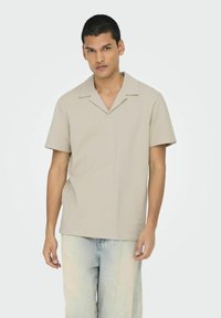 Camicia beige a maniche corte con colletto, tessuto testurizzato e vestibilità rilassata, abbinata a pantaloni in denim di colore chiaro.