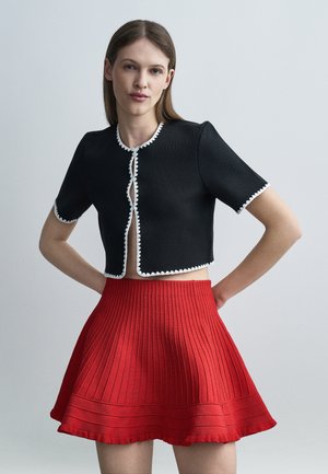 Kobieta o prostych brązowych włosach, ubrana w czarny krótki top z białą lamówką i czerwoną plisowaną spódnicę z wysokim stanem, stojąca z rękami na biodrach.