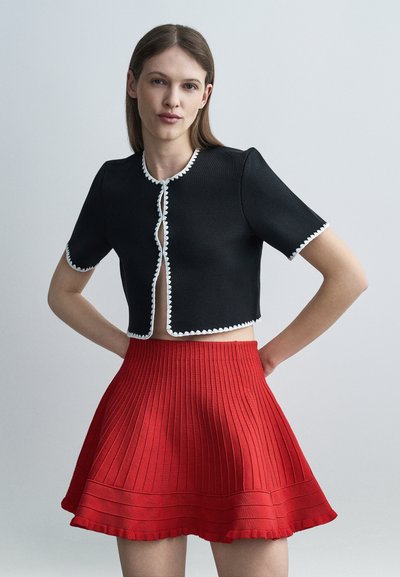 Femme aux cheveux bruns raides portant un haut court noir à bord blanc et une jupe plissée rouge taille haute, debout les mains sur les hanches.