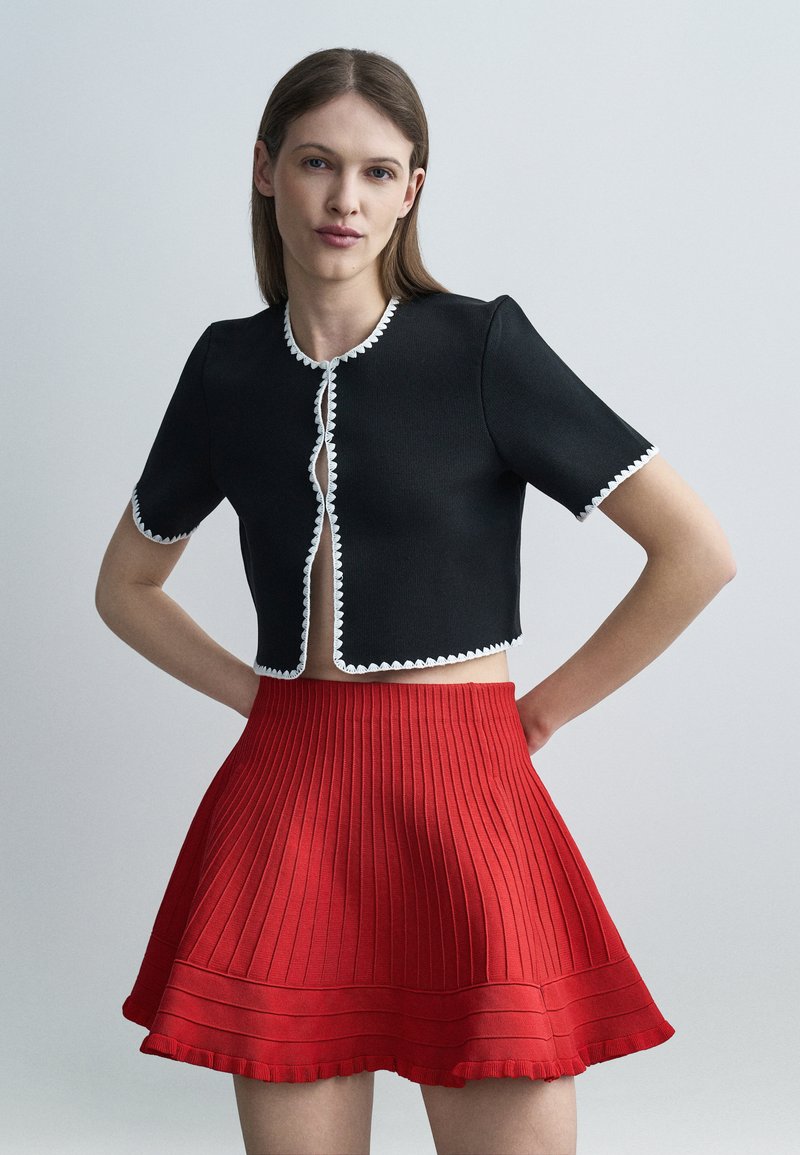 Γυναίκα με ίσια καστανά μαλλιά, που φοράει μαύρο cropped τοπ με λευκή επένδυση και κόκκινη πλισέ φούστα μέσης, στέκεται με τα χέρια στη μέση.