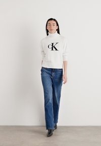 Hvit strikket genser med høy hals, som har en svart "CK" logo. Kombinert med blå straight-leg jeans og svarte ankelstøvler.