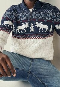 Pull-over en tricot avec un motif bleu marine et blanc représentant des élans et des arbres. Section inférieure en tricot torsadé blanc ; le col zippé ajoute de la fonctionnalité.