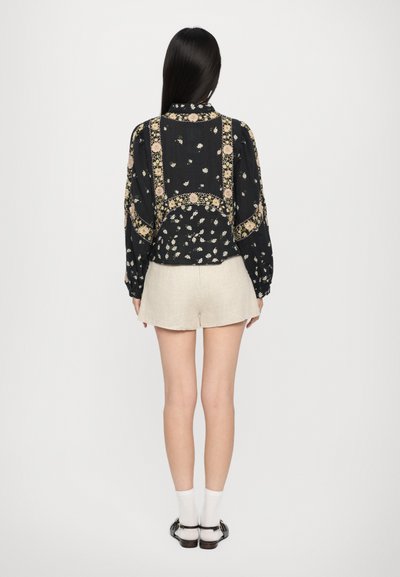 Blusa nera a fiori con accenti gialli e un design con colletto, abbinata a pantaloni corti di lino beige. Dettagli visibili sulla schiena e maniche arricciate.