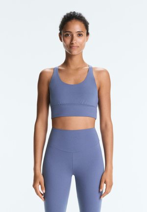 Ženska stoji obrnjena naprej, oblečena v usklajen modri športni crop top in visokim pasom legice na enobarvni podlagi.