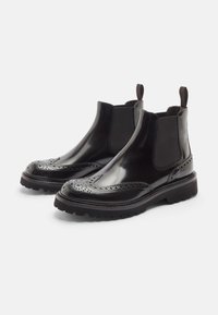 Botas Chelsea de cuero negro con acabado pulido, detalles de brogue en la punta, paneles laterales elásticos y suela de goma texturizada.