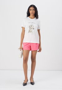 T-shirt en coton blanc avec un graphique de chien tacheté, associé à un short rose. Le mannequin tient un petit sac à main blanc et porte des ballerines noires.