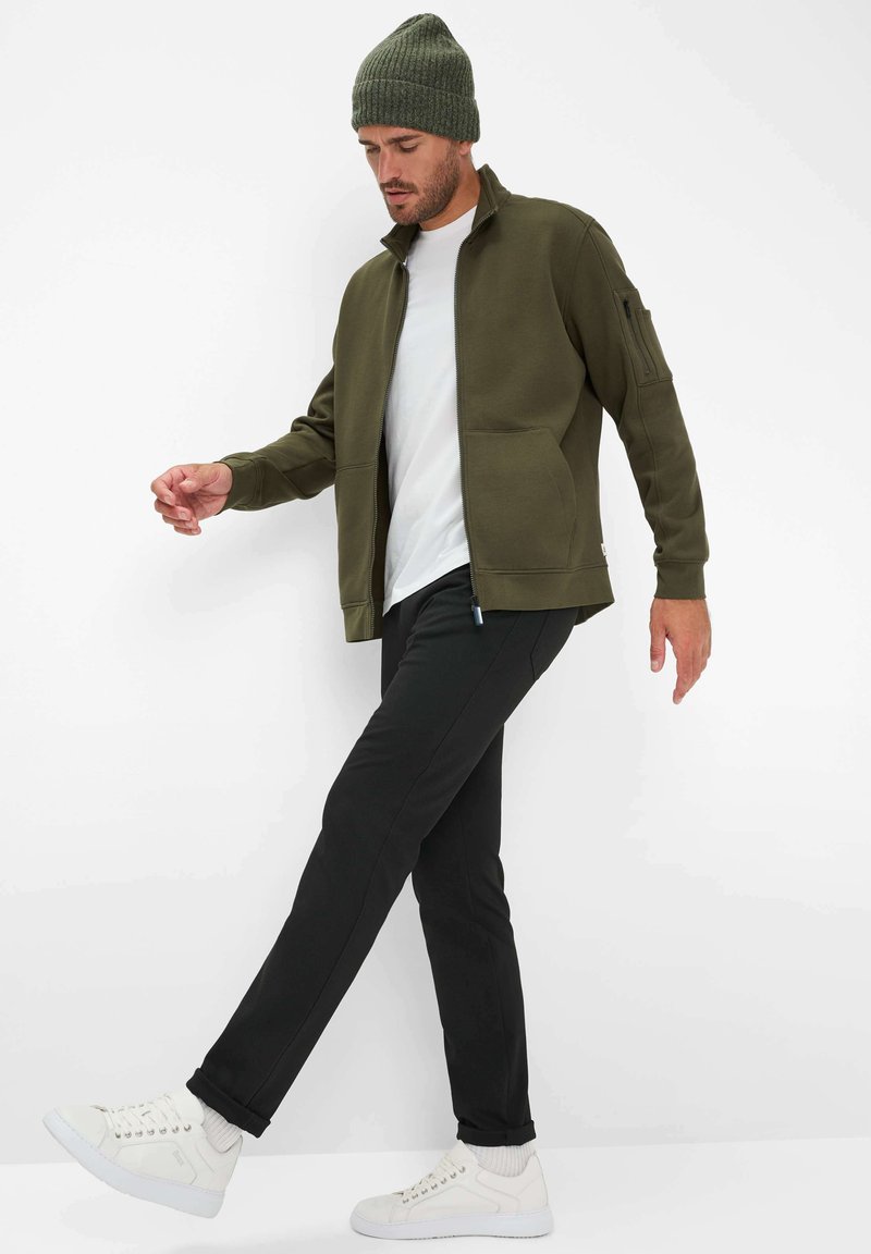 Giacca verde oliva con zip e colletto alto, t-shirt bianca, pantaloni neri e sneakers bianche. La giacca ha una tasca sulla spalla ed è realizzata in un tessuto morbido.