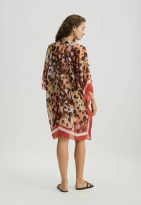 Chiffon strandbedekking met een luipaardpatroon in bruin en oranje tinten, met rode en witte bies langs de zoom en mouwen.