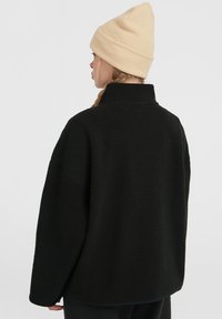 Black fleece jacket with a high collar and oversized fit. The fabric has a textured surface. A beige knit beanie is worn on the head.