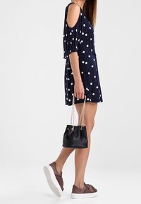 Abito navy a pois con spalle scoperte, borsa nera a secchiello con tracolla in catena e sneakers slip-on in suede marrone con suole bianche.