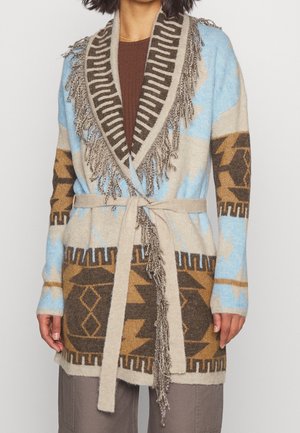 Frau trägt einen gestrickten, mit Gürtel versehenen Cardigan mit blauen, braunen und beigen geometrischen Mustern und einem Fransen-Schalkragen über einem braunen Top.