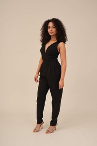Svart jumpsuit med dyp V-utringning, tettsittende liv og plissert midje. Stilet med beige hæler med stropper, på en nøytral bakgrunn.