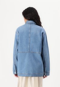 Lys blå denimjakke med krage, lange ermer og en avslappet passform. Har synlige sømmer og knappdetaljer.