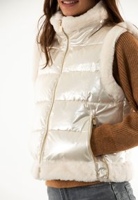 Gilet smanicato effetto puff in materiale metallico lucido color crema, con un morbido colletto e orlo in pelliccia sintetica, zip dorata e dettagli di cucitura.