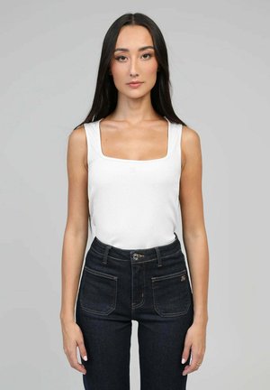 Femme aux longs cheveux foncés portant un haut blanc sans manches à col carré et un jean foncé taille haute, debout devant un fond uni.