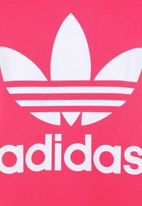 Rosa adidas-textil med en stor vit trebladig logotyp och djärv vit märkning nedanför. Slät textur med en livfull färgkontrast.
