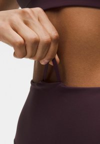 Mörklila sportstopp med en mjuk, elastisk textur. En hand drar i en tygrem från sidan, vilket betonar plaggets design.