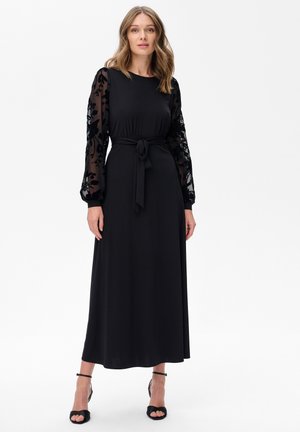 Czarna sukienka maxi z długimi koronkowymi rękawami, dopasowanym topem i pasującym paskiem w talii. Gładki materiał, elegancki design i sandały na odkrytej pięcie.