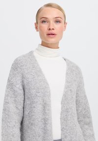 Cardigan grigio chiaro, fuzzy, con frontale aperto e spalle cadenti, indossato sopra un dolcevita bianco. Texture morbida con dettagli di design minimi.