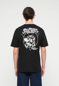 T-shirt de algodão preto com impressão gráfica branca nas costas, apresentando um design de lobo e o texto "O Saloon do Lobo Solitário Mais Solitário."