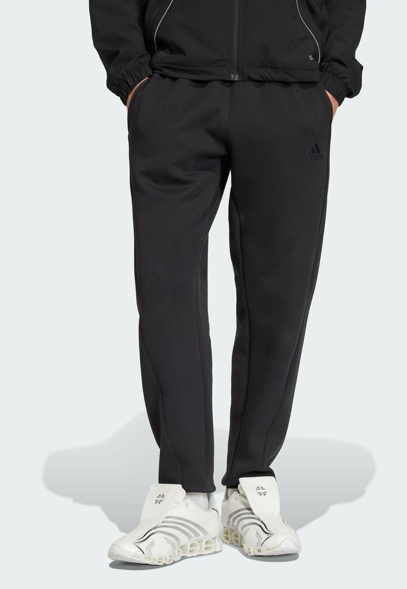 Persona che indossa pantaloni da tuta neri Adidas con le mani nelle tasche e sneakers bianche Adidas con un design di suola unico.